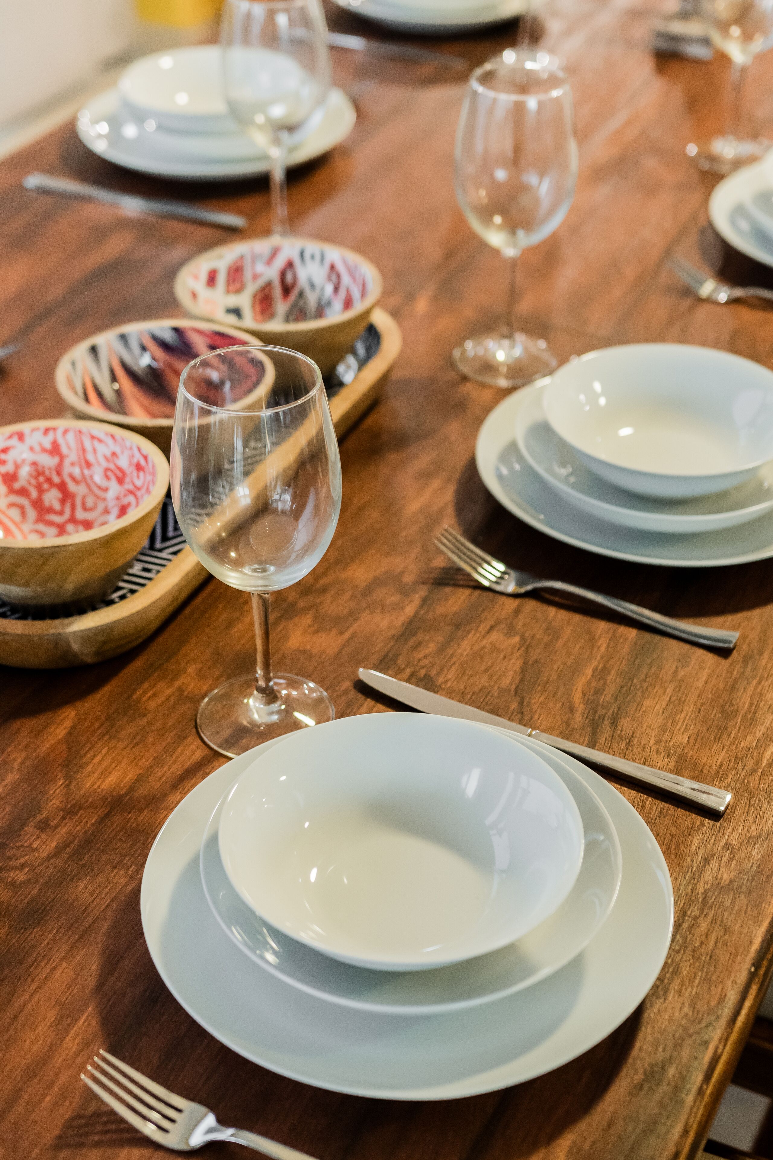 A dining table set with white plates, bowls, and wine glasses, with a wooden table and a fork on the left side.