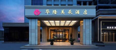 A hotel entrance with a modern facade, a prominent logo above the entrance, and a well-lit interior visible through the glass doors.