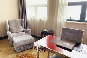 Habitación de hotel con un sillón tapizado de gris, una mesita redonda con una caja de pañuelos y una ventana con cortinas de gasa.