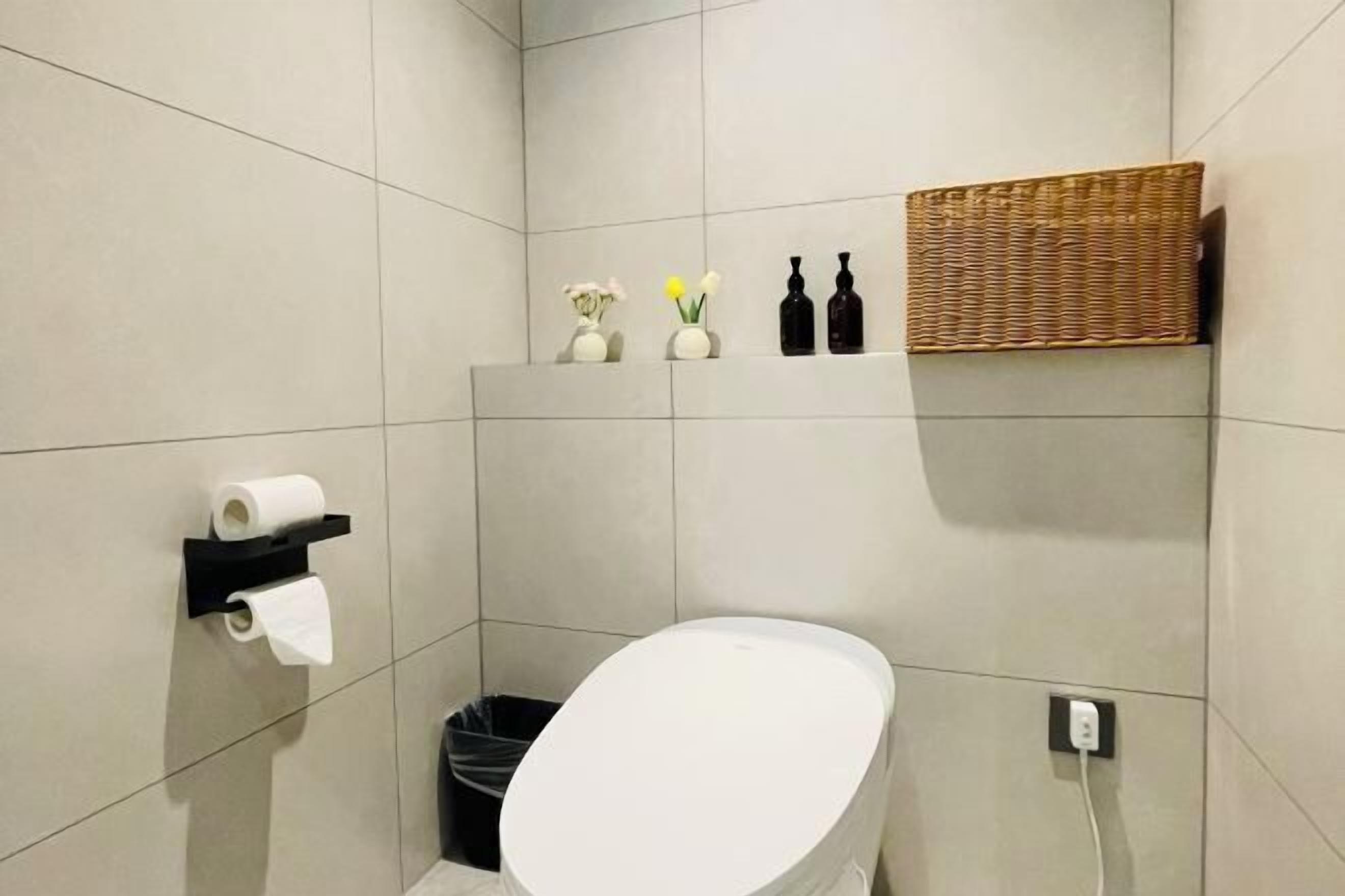 A modern bathroom with a white toilet, a black trash bin, a shelf with toiletries, and a wicker basket.