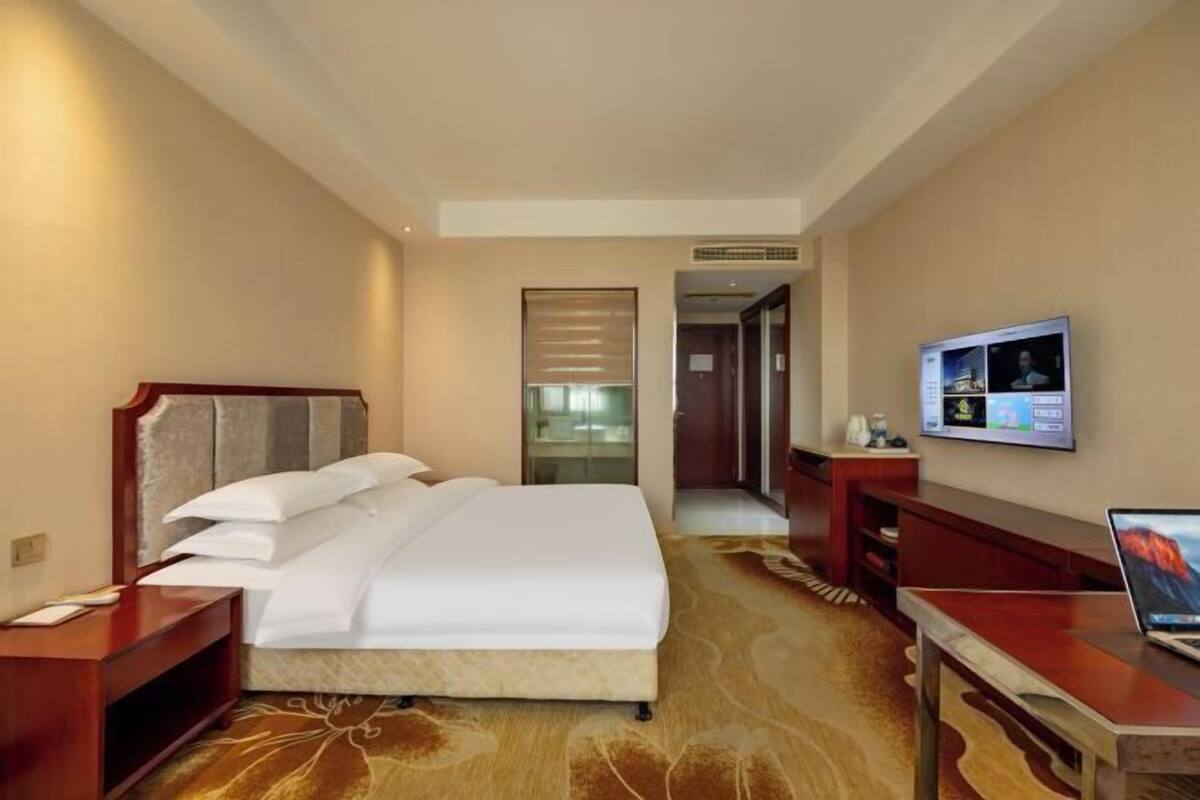 A hotel room with a bed, a desk with a laptop, a TV, and a carpeted floor.