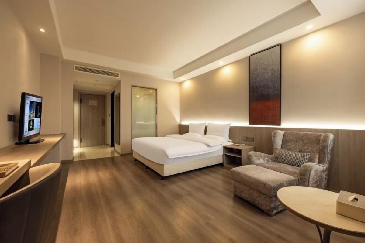 A modern hotel room with a bed, a desk with a computer, a chair, and a small table.
