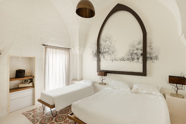 A bedroom with two beds, a wooden shelf with laundry, a window with curtains, and a wall-mounted mirror. - Al Palazzo La Dimora (Fasano)