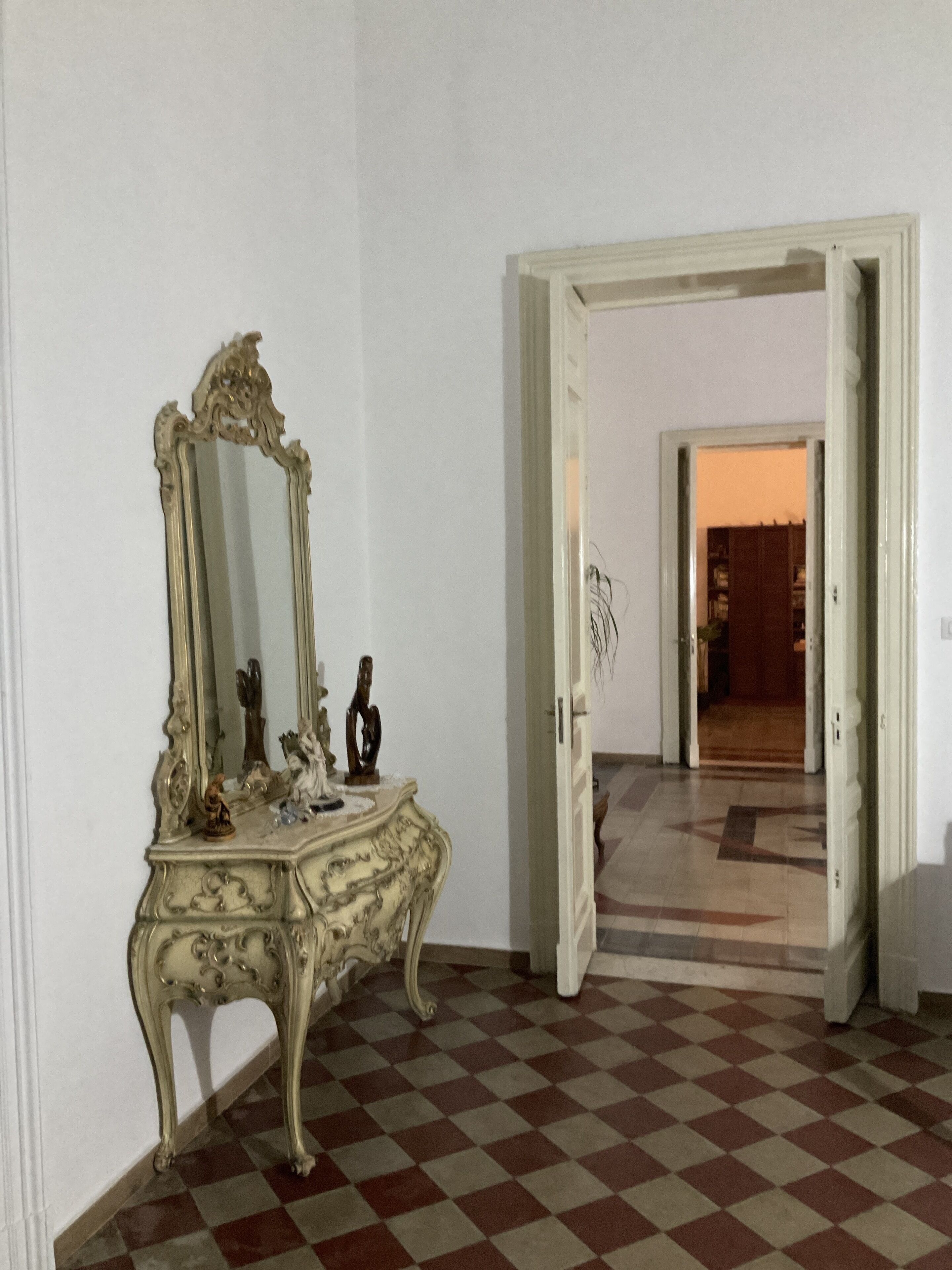 Um quarto com chão quadriculado, um espelho grande e ornamentado e uma porta que dá para outro quarto.