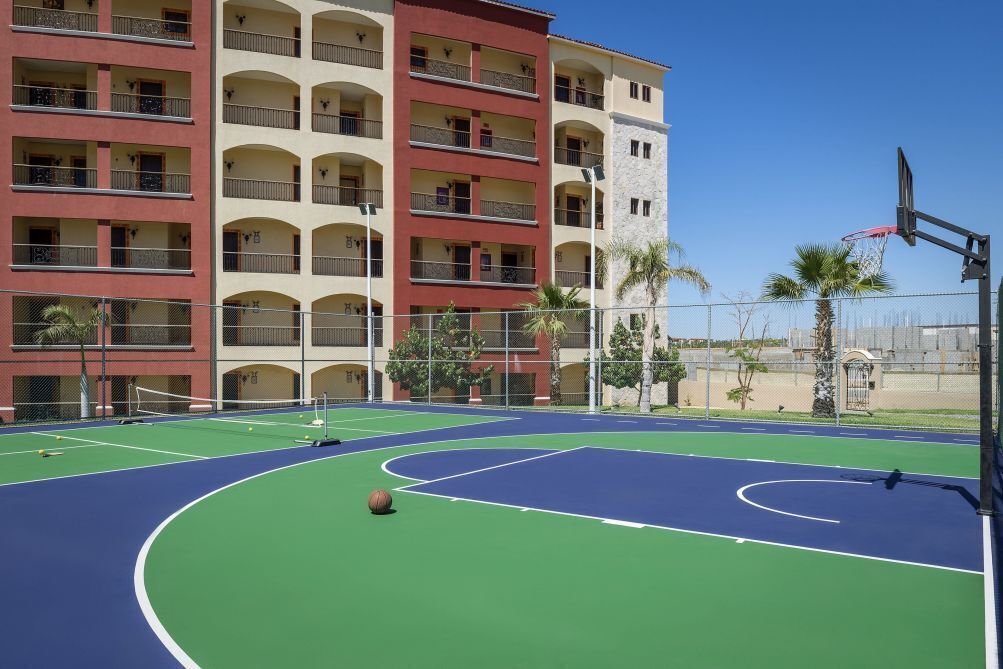 An outdoor basketball court with a blue and green surface, surrounded by a fence and palm trees.
