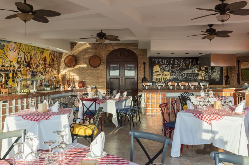 A restaurant with multiple tables set for dining, a bar area with various bottles, and a mural depicting a cityscape.