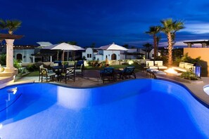 A swimming pool with lounge chairs and umbrellas, surrounded by palm trees and a building with arched windows. - Experience the Ultimate in Comfort & Convenience: 3BR Condo with Pool (Cabo San Lucas)