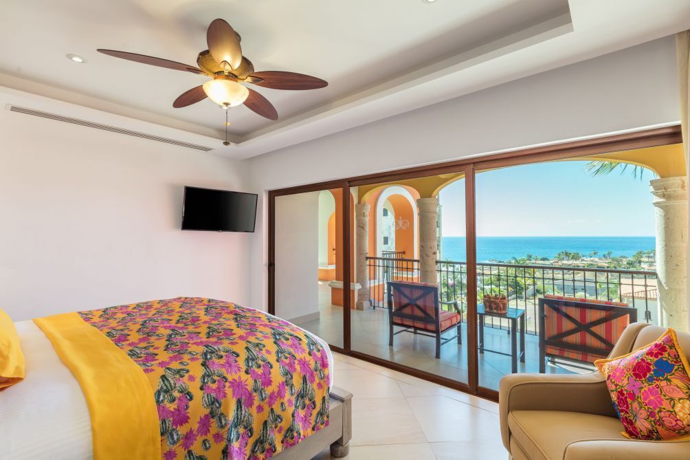 Une chambre avec un lit, un ventilateur de plafond, une télévision et un balcon offrant une vue sur l’océan.