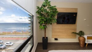 A modern bedroom with a large window, a potted plant, a painting, and a view of the sea.