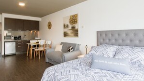 A modern apartment with a kitchenette, a dining area, and a living room with a grey sofa and a bed with patterned bedding.