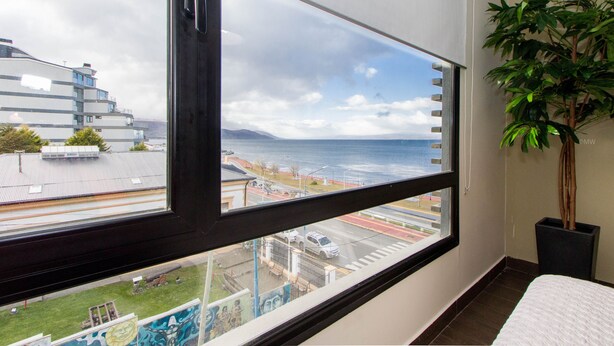 A room with a large window offering a view of a coastal area, a potted plant, and a bed.