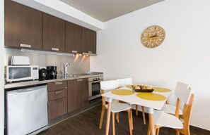 Une cuisine moderne avec des meubles en bois foncé, un réfrigérateur blanc, un four à micro-ondes, une machine à café et une cuisinière. On y trouve une table à manger avec des sets de table jaunes et des chaises blanches. Une horloge murale ronde est visible au mur.