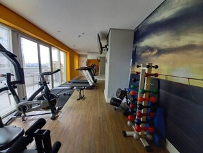 Uma academia com equipamentos de cardio, estações de musculação e uma grande janela com vista para a cidade.