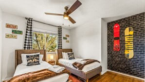 A hotel room with two beds, a ceiling fan, a window with curtains, a brick wall with a snowboard, and a wooden floor. - The Mountain Retreat: Steps to Palmer Park, Central COS, Sleeps 8 (Colorado Springs)