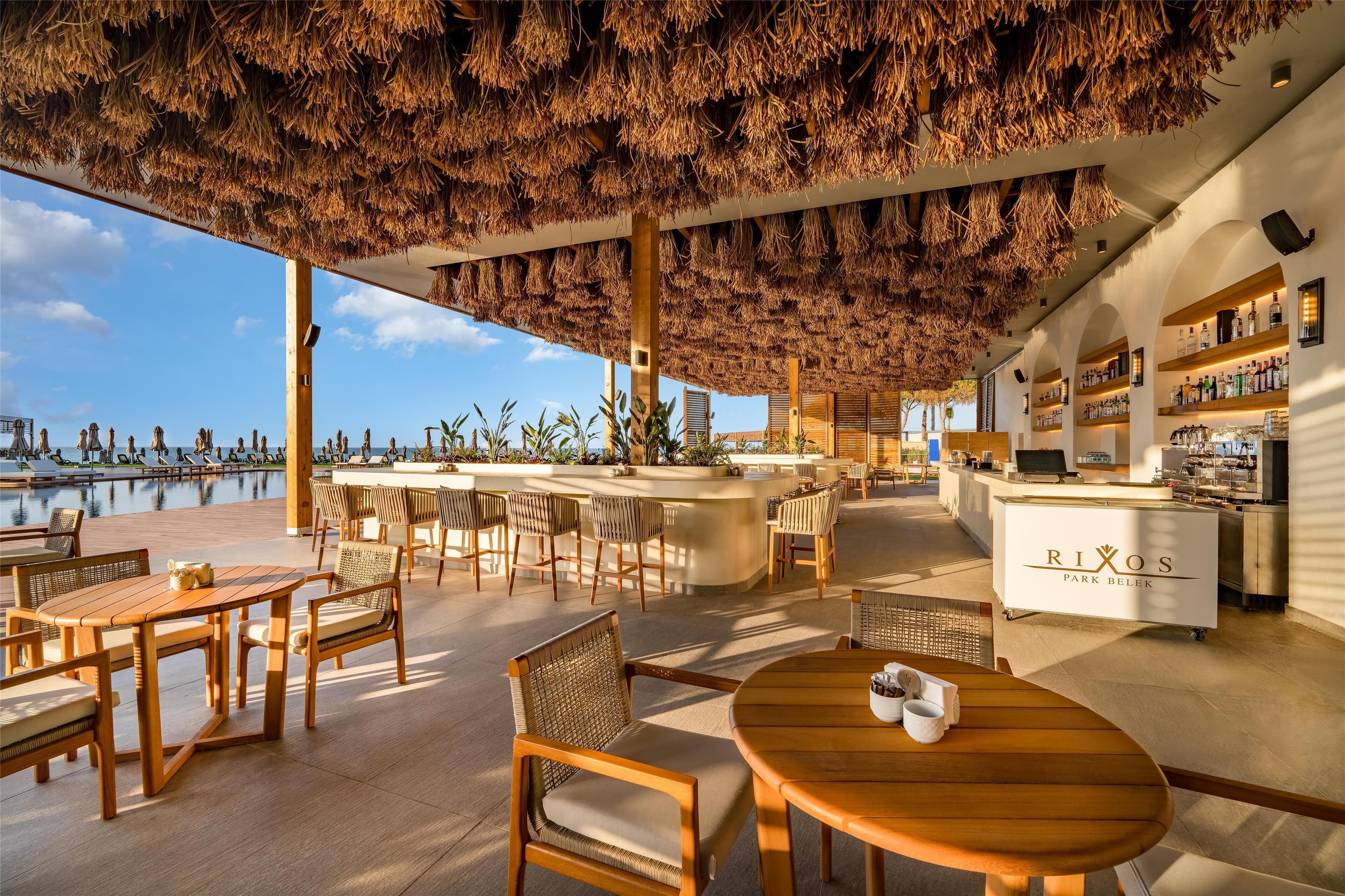 An outdoor bar area with wooden furniture and a thatched roof, overlooking a body of water.