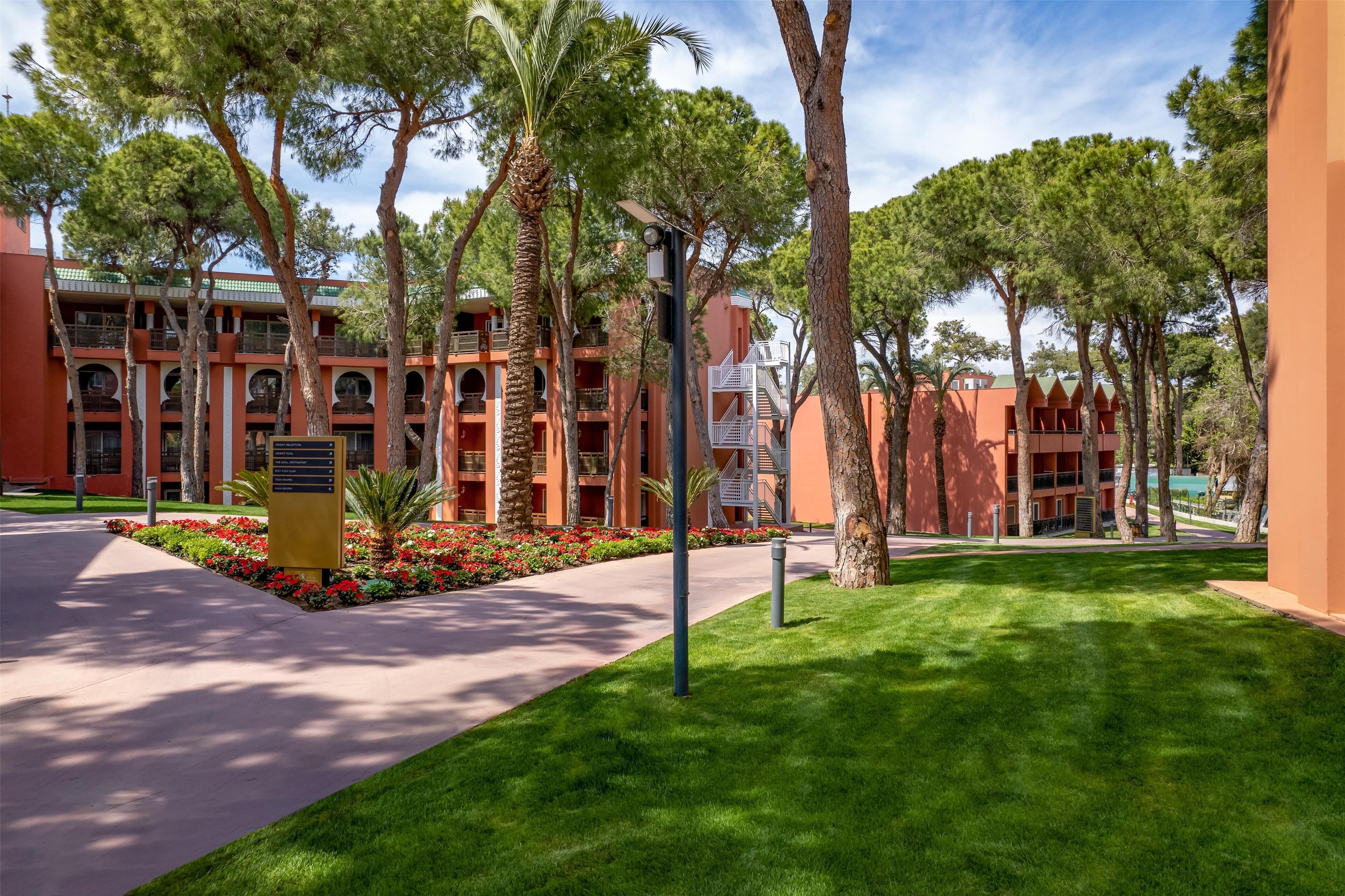 Un patio de hotel con un camino central, rodeado de palmeras y plantas de flores rojas.
