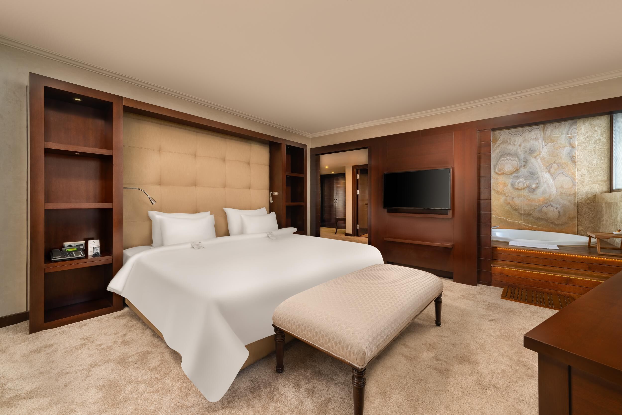 A spacious bedroom with a large bed, a bench, a TV, and a bathroom with a marble wall.
