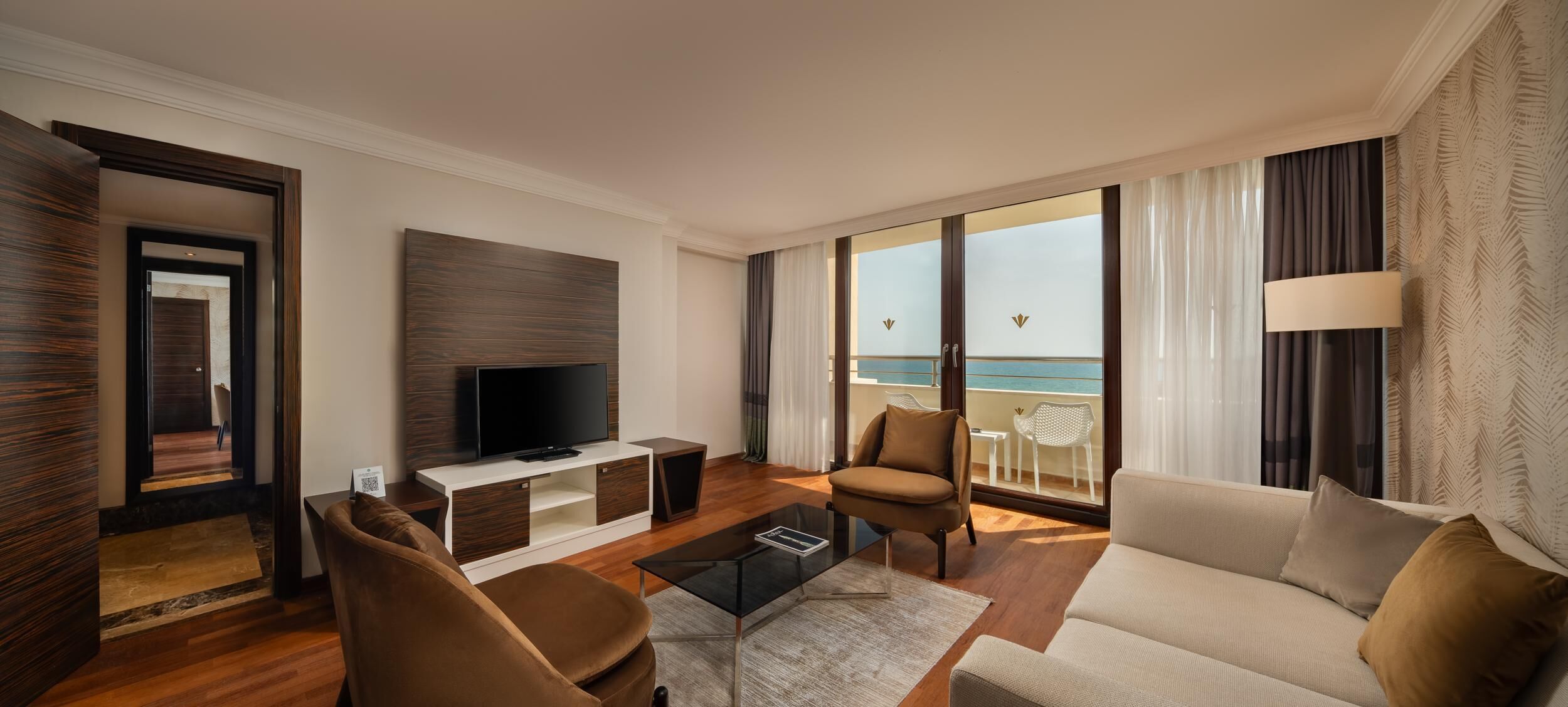 A modern living room with a TV, sofa, and armchair, a balcony with a view of the sea, and a wooden floor.
