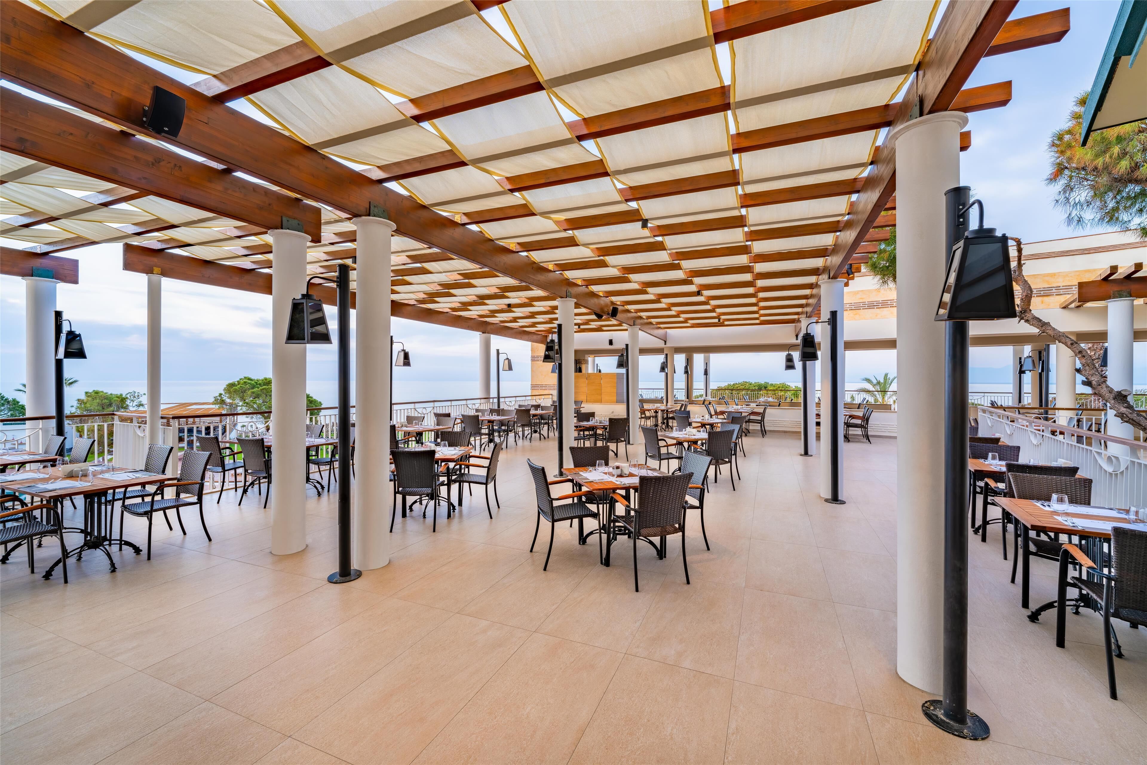 An outdoor dining area with multiple tables and chairs, a covered roof structure, and a view of the sea.