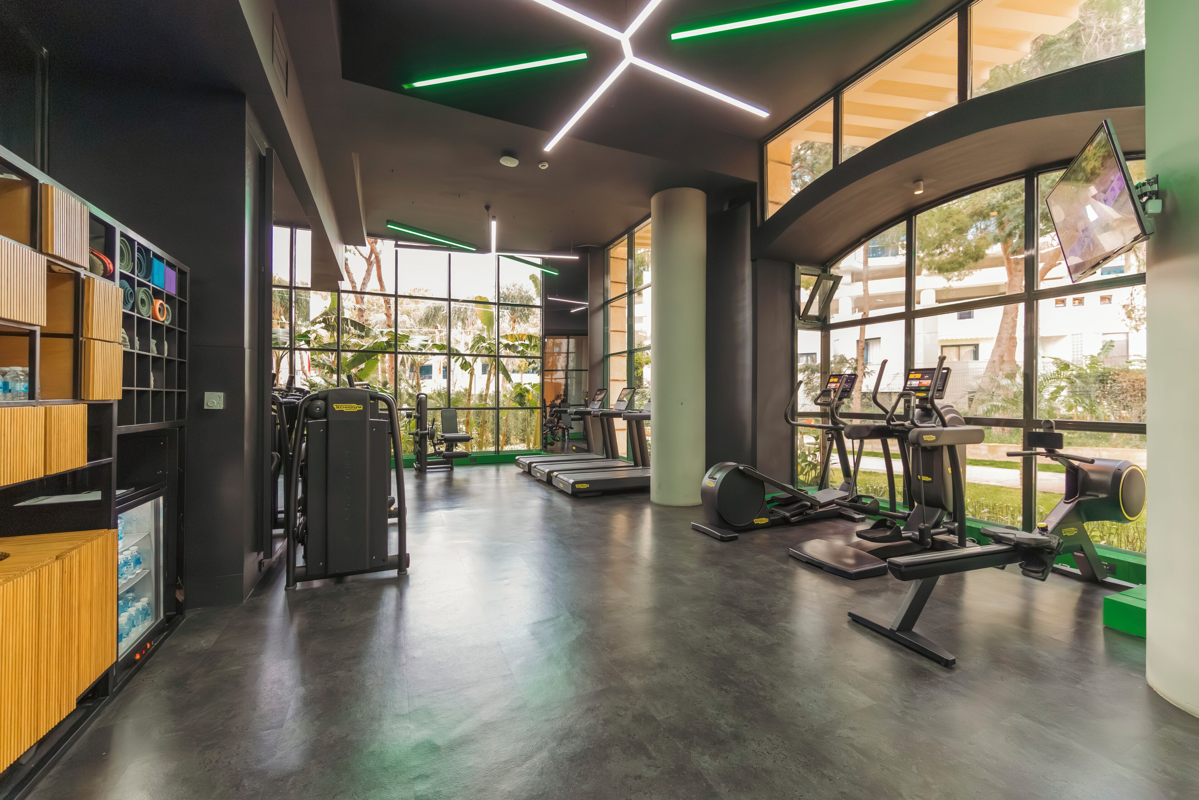 A modern gym with exercise equipment, large windows, and a wooden wall with shelves.