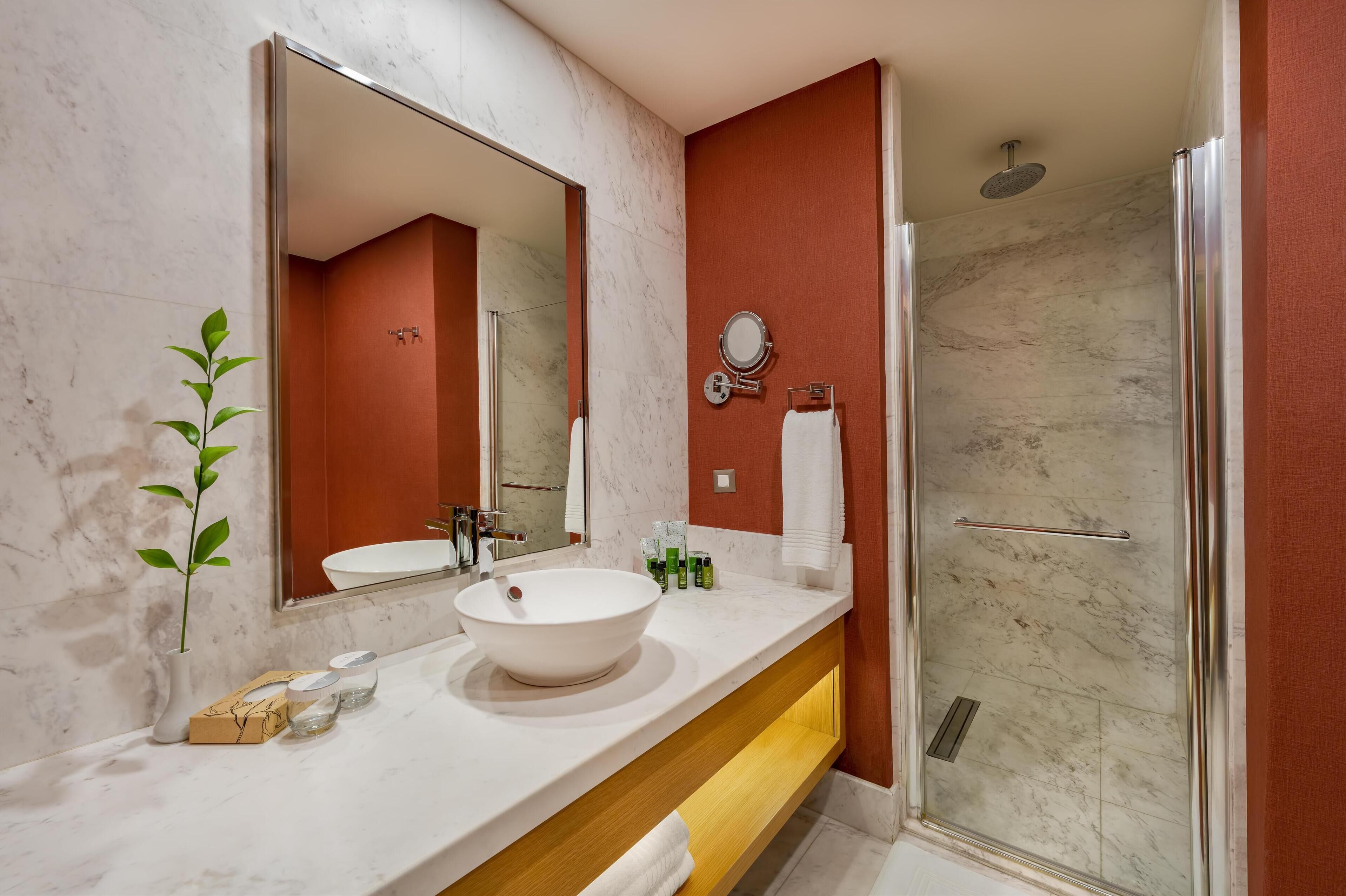 A modern bathroom with a large mirror, a white sink, a glass shower enclosure, and a potted plant.