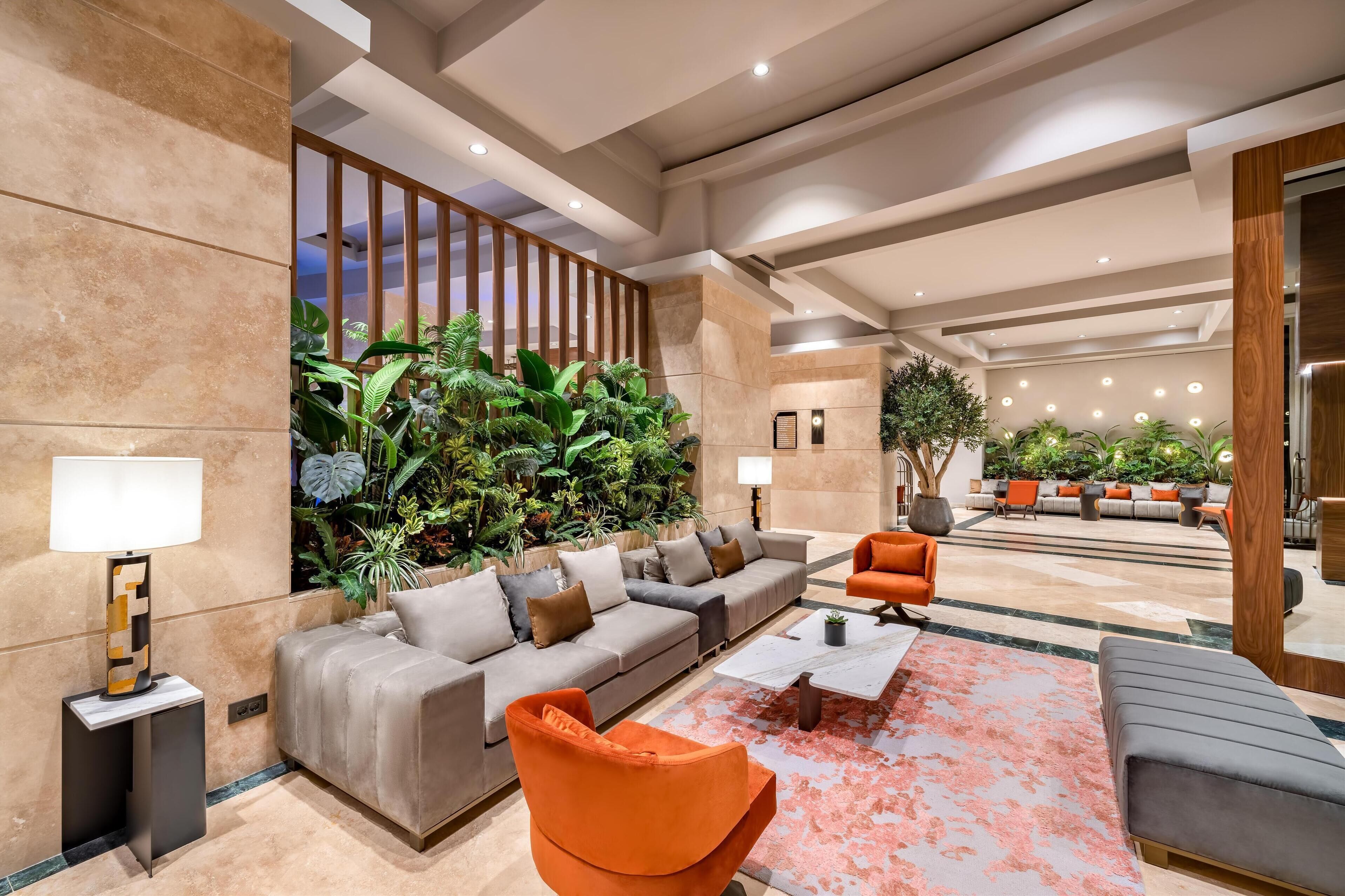 A modern lobby with a sofa, armchairs, and a coffee table. There are plants and a seating area in the background.
