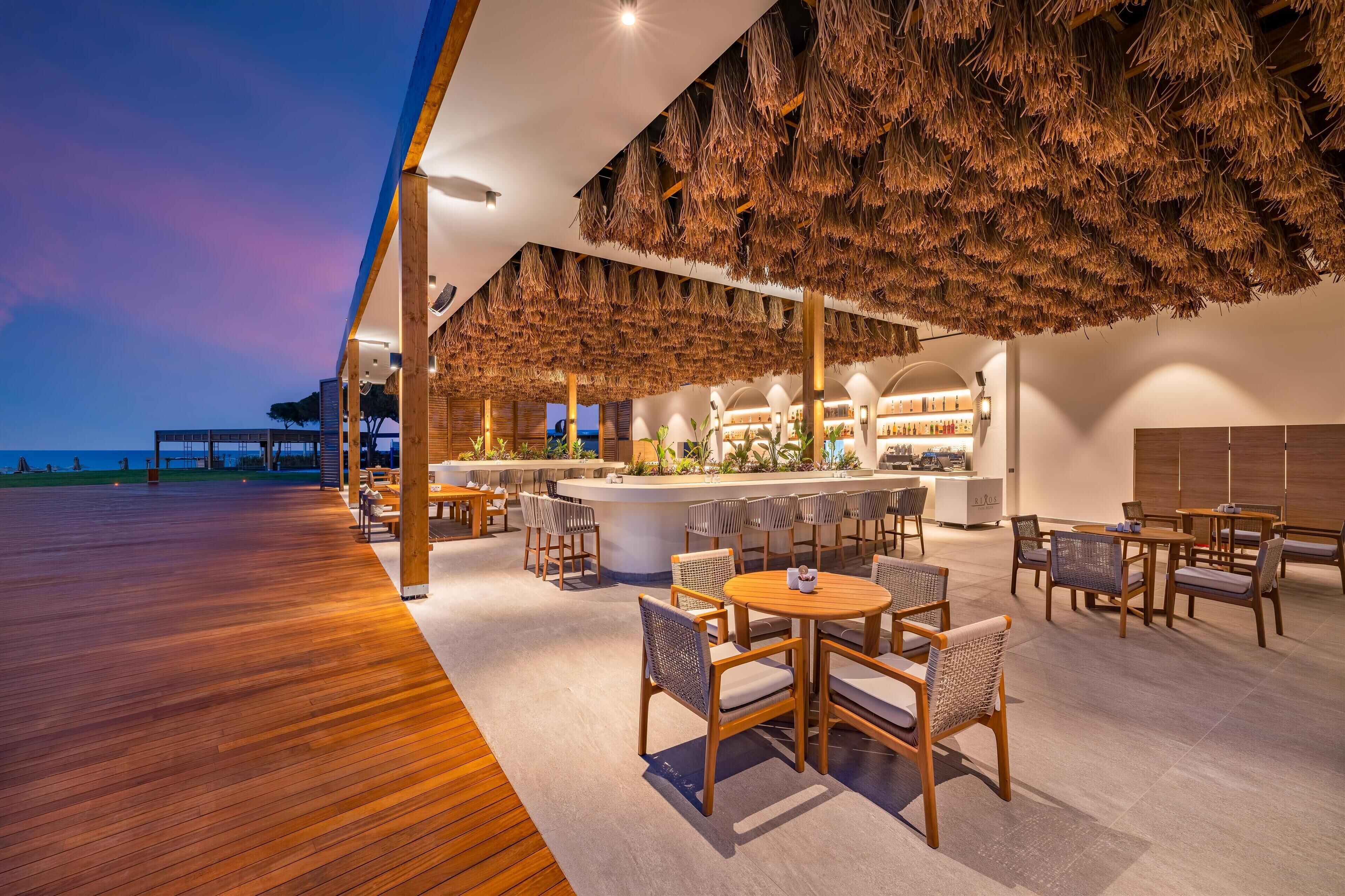 A modern outdoor dining area with wooden flooring, thatched ceiling, and a bar with seating.
