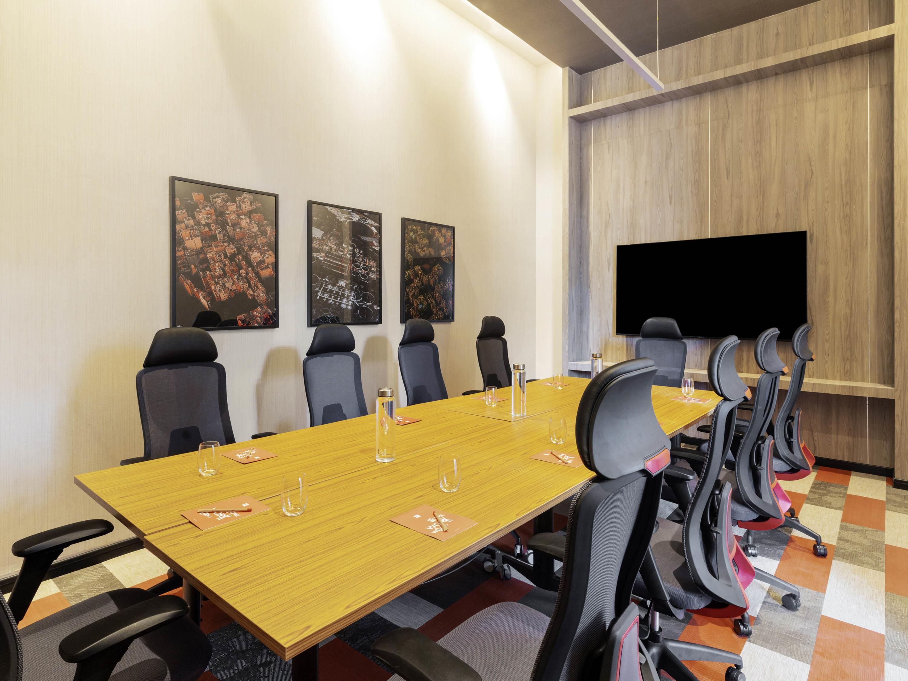 A conference room with a long table, multiple chairs, a flat-screen TV, and three framed pictures on the wall.