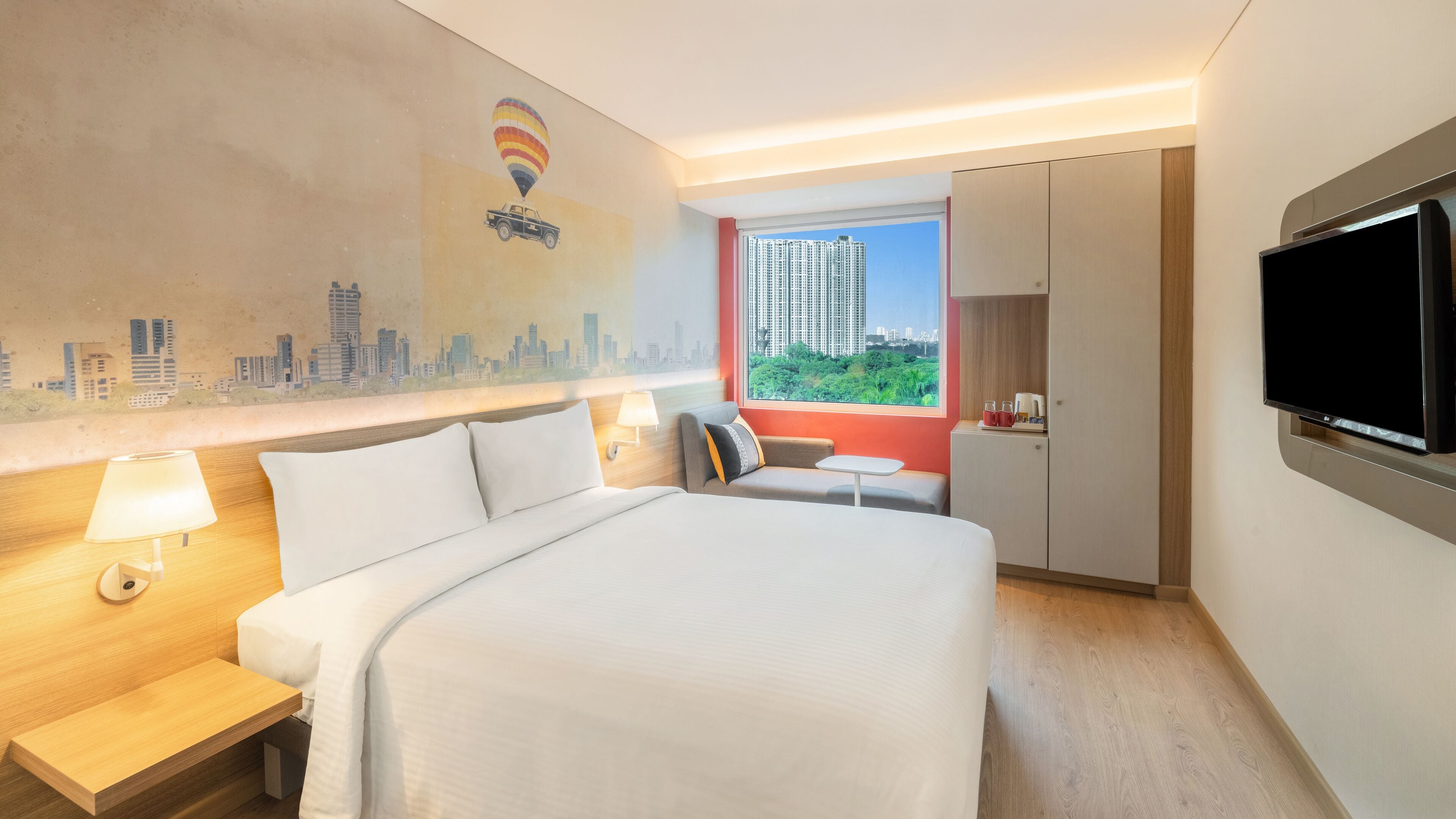 A hotel room with a large bed, a desk, a chair, a TV, and a window with a cityscape view.