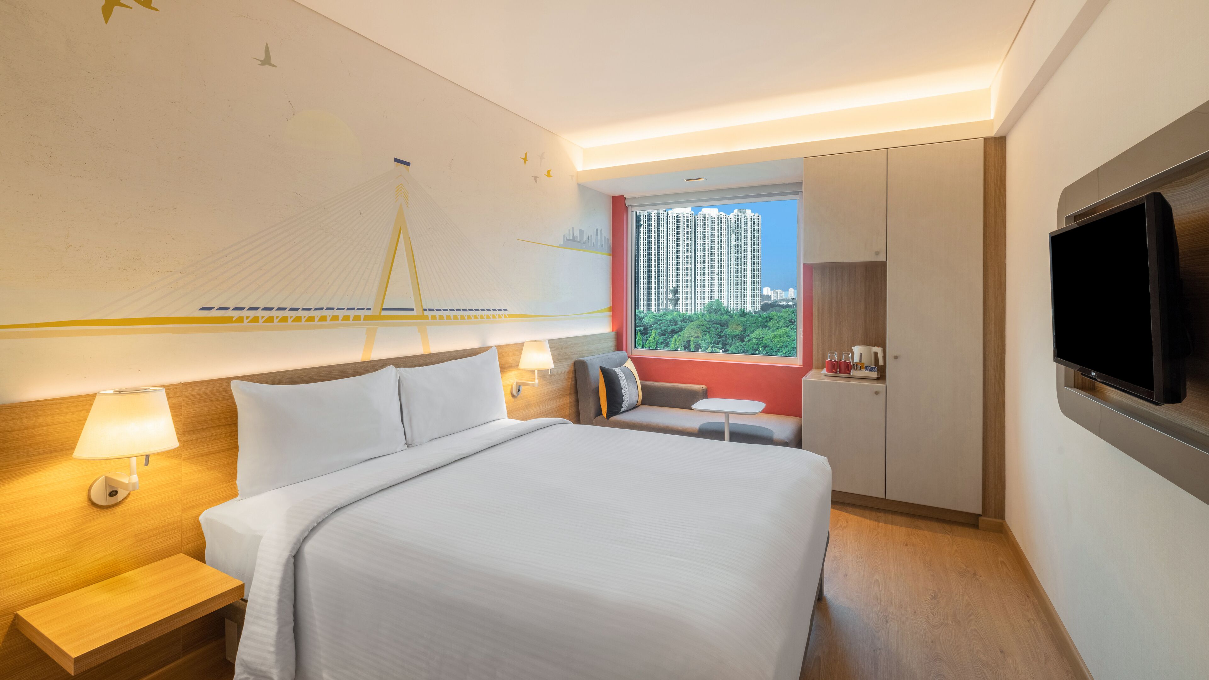 Ein Hotelzimmer mit einem großen Bett, einem Fernseher, einem Schreibtisch mit Lampe, einem kleinen Tisch und einem Fenster mit Blick auf die Stadt.