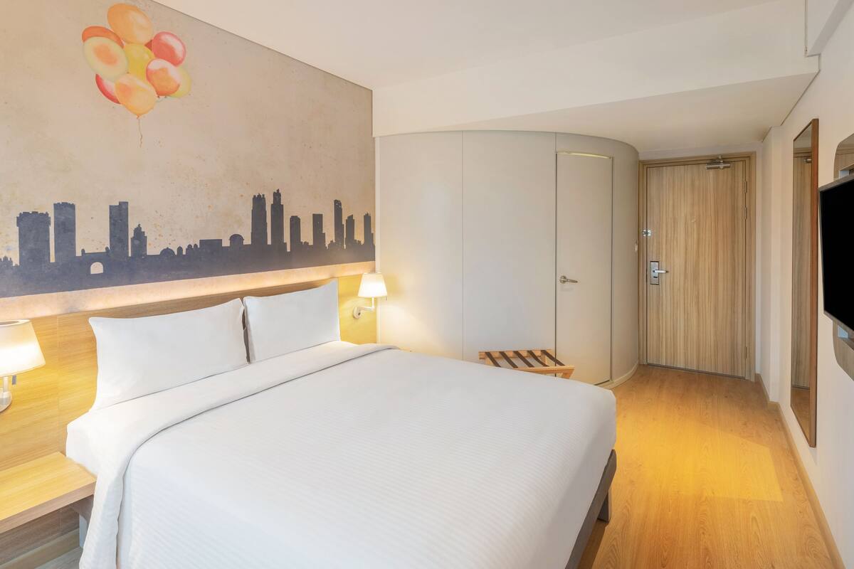 A hotel room with a bed, two bedside lamps, a cityscape mural, a wardrobe, a TV, and a wooden bench.