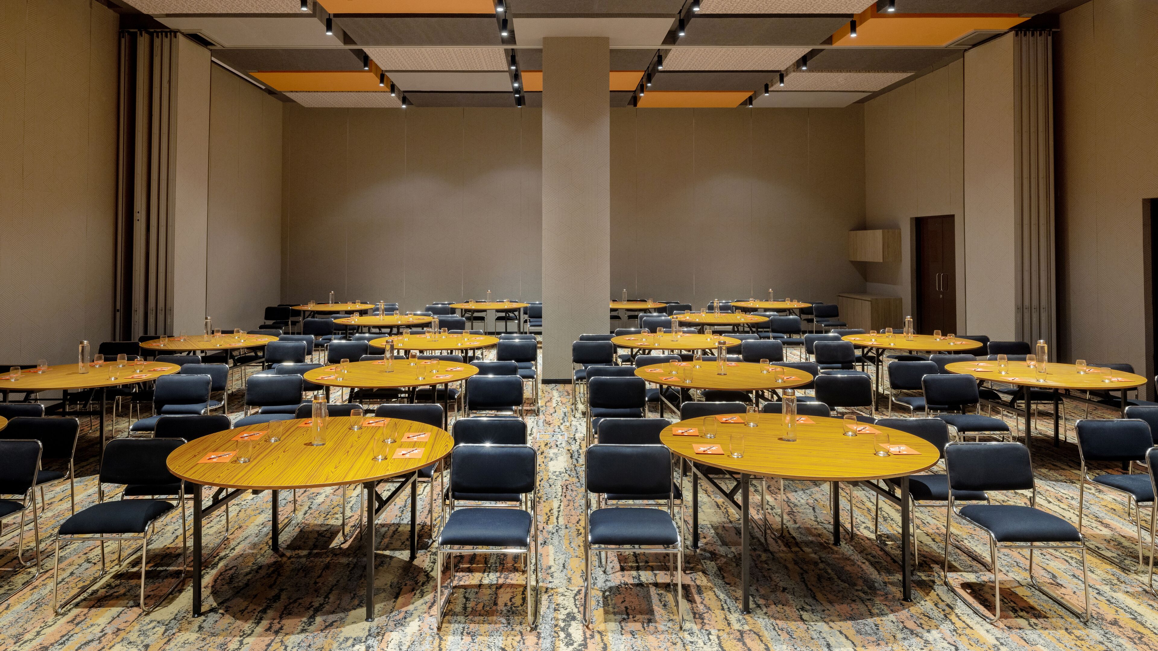 A conference room with multiple round tables and chairs, carpeted flooring, and a high ceiling with recessed lighting.