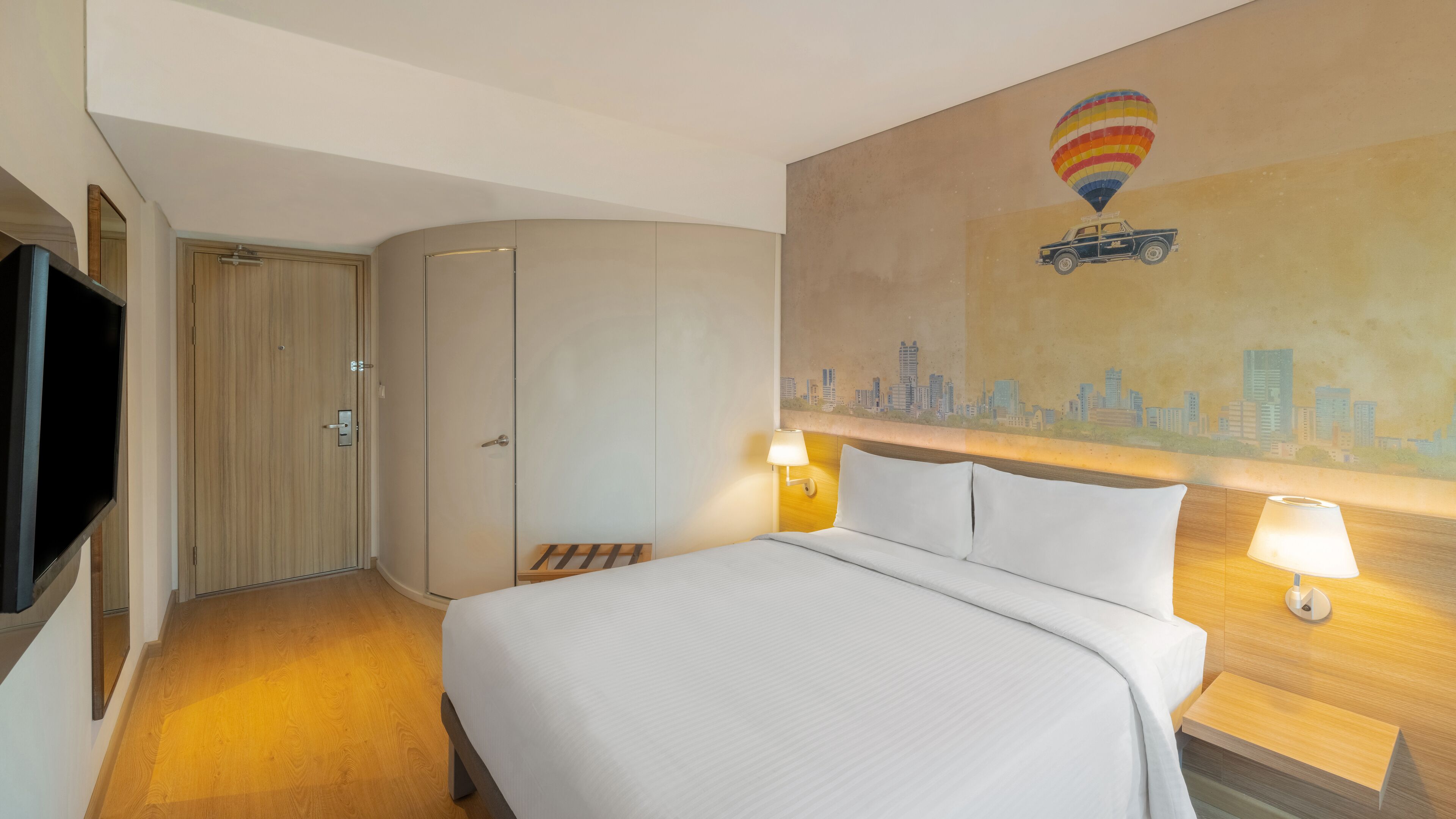 A hotel room with a bed, a TV, a wardrobe, and a painting of a hot air balloon over a cityscape.