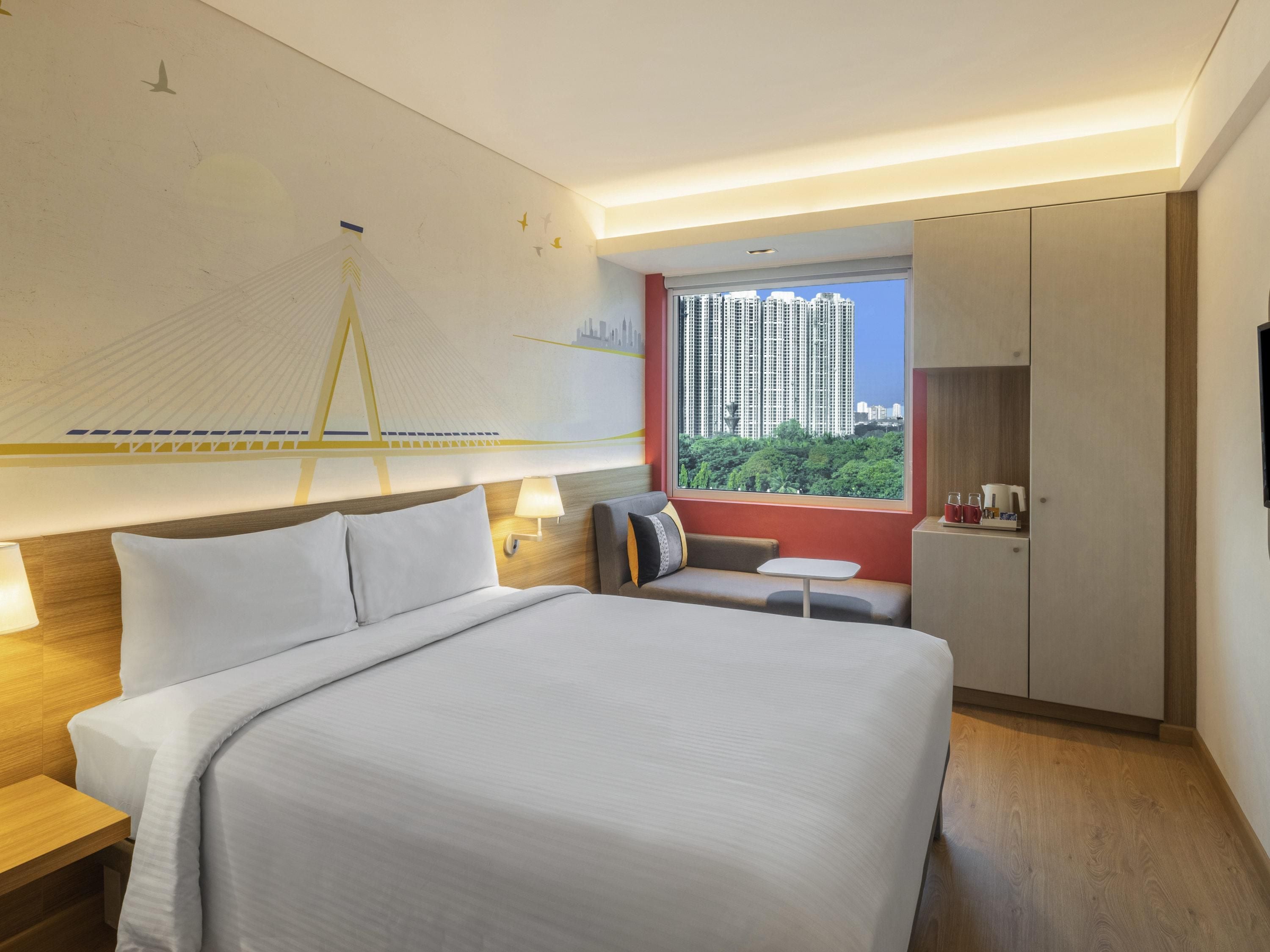 A hotel room with a bed, bedside table, lamp, and a view of high-rise buildings.