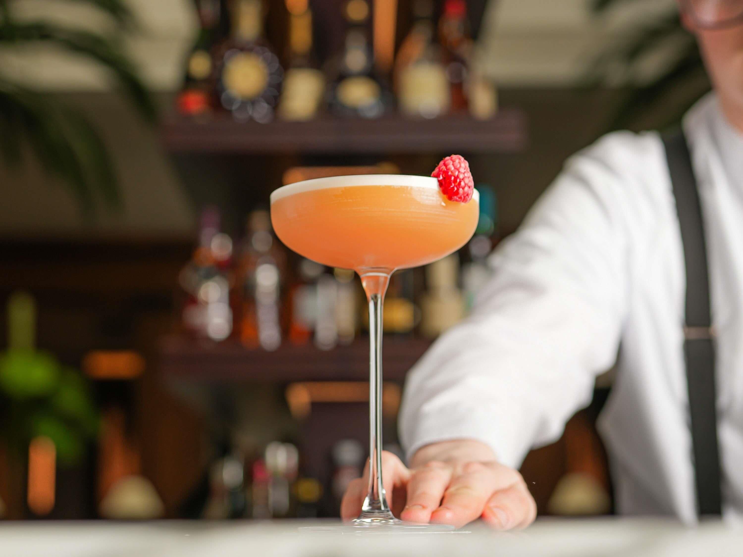A cocktail in a martini glass with a garnish, a person in a white shirt, and a blurred background with shelves of bottles.