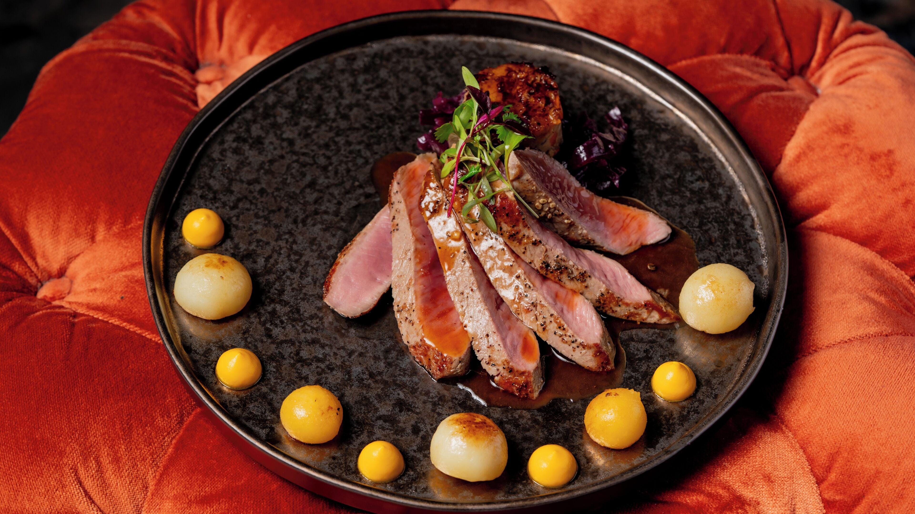 A plate of sliced meat with a garnish, surrounded by small round items, on a dark plate.