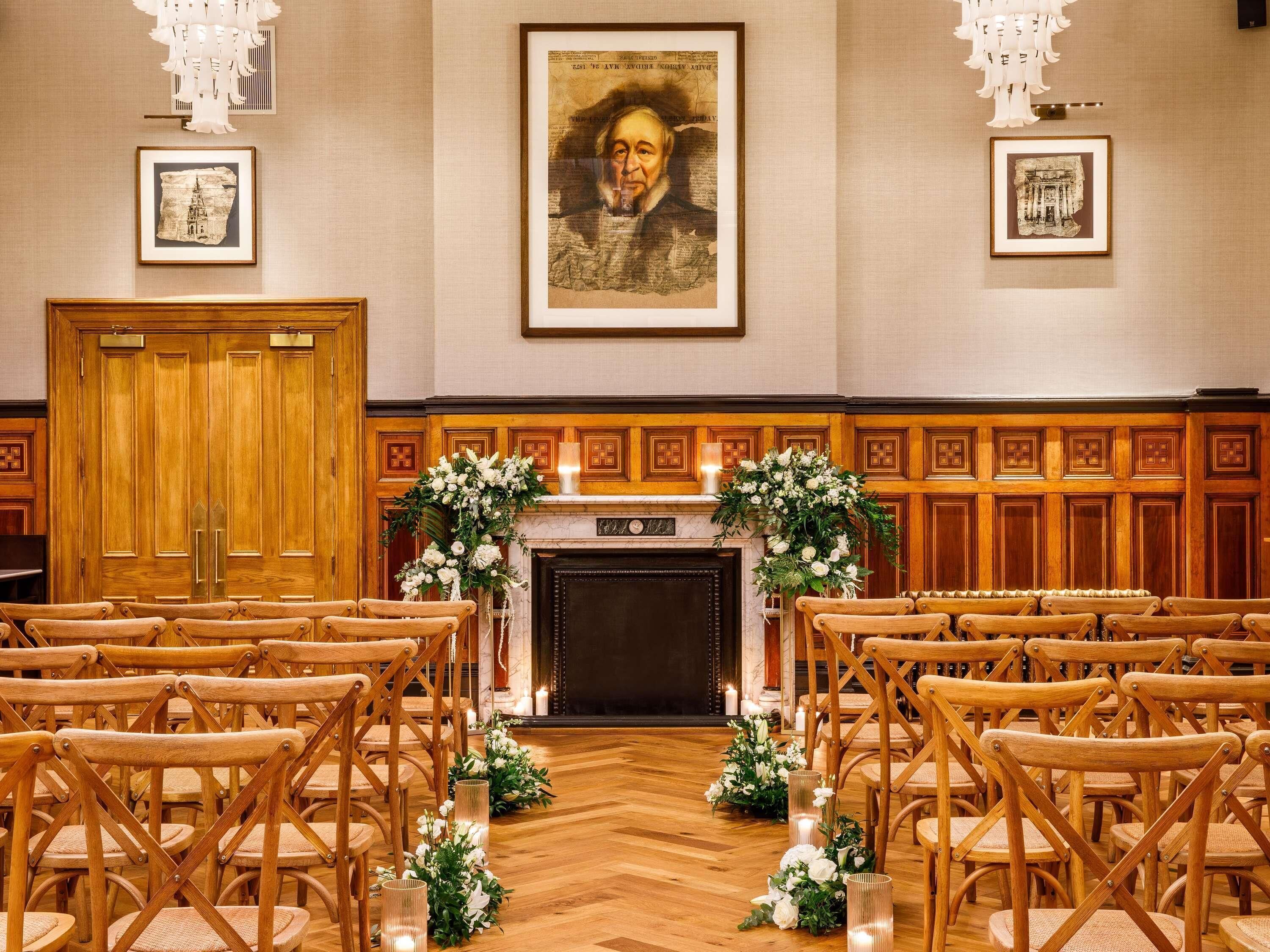 A formal room with wooden chairs arranged in rows, a fireplace with floral decorations, and framed pictures on the walls.