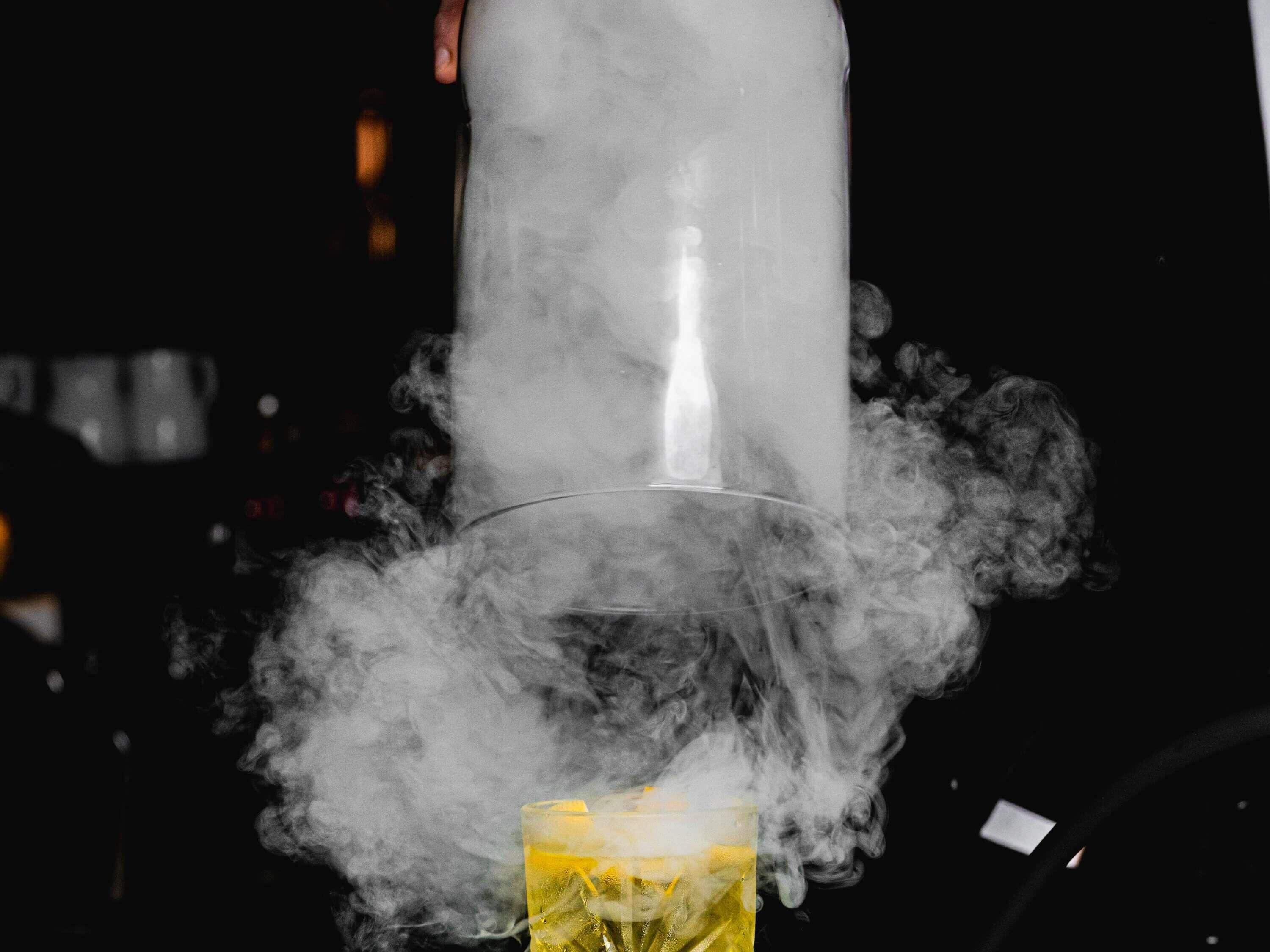 A person pouring a drink from a shaker into a glass, surrounded by smoke.