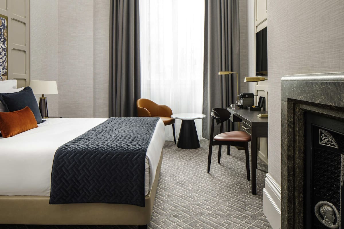 A hotel room with a bed, a desk, a chair, a fireplace, and a view of the outdoors.