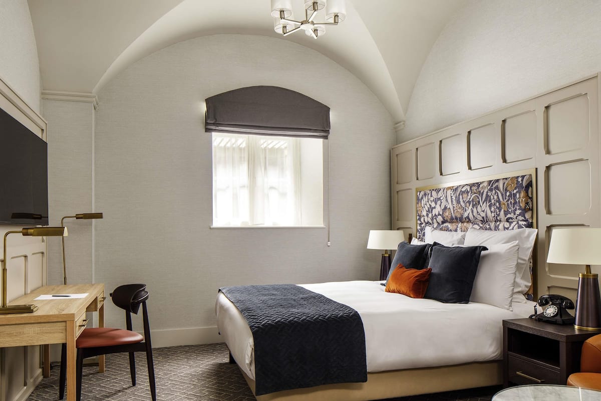 A hotel room with a bed, bedside tables, a desk, a chair, a TV, and a window with curtains.