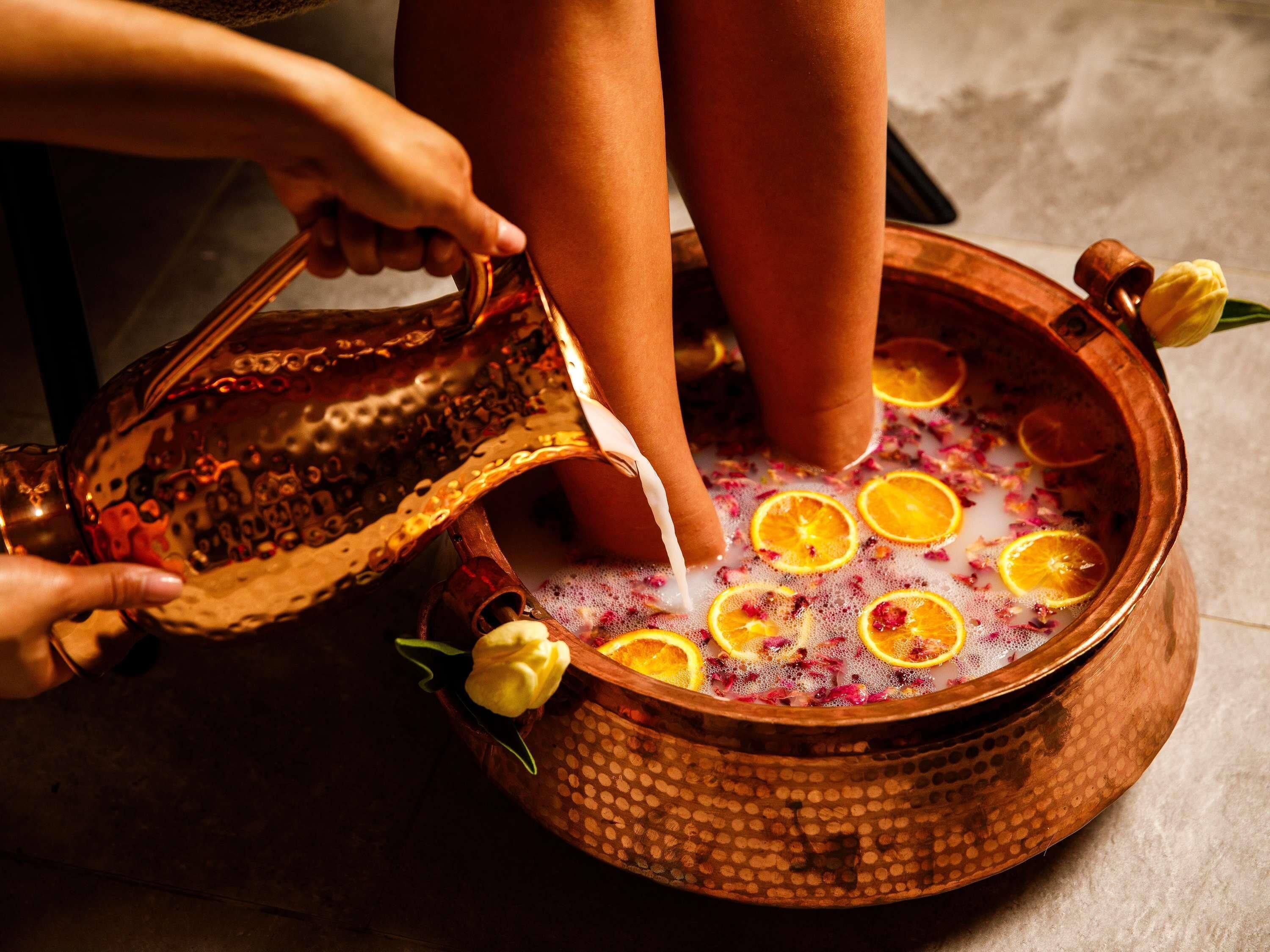 A copper basin filled with a bubbling liquid, garnished with orange slices and flowers, with a person pouring from a patterned bowl.