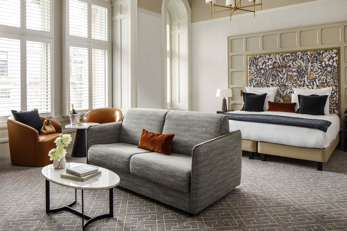 A hotel room with a grey sofa, a brown armchair, a round coffee table, a bed with a patterned headboard, and a chandelier.