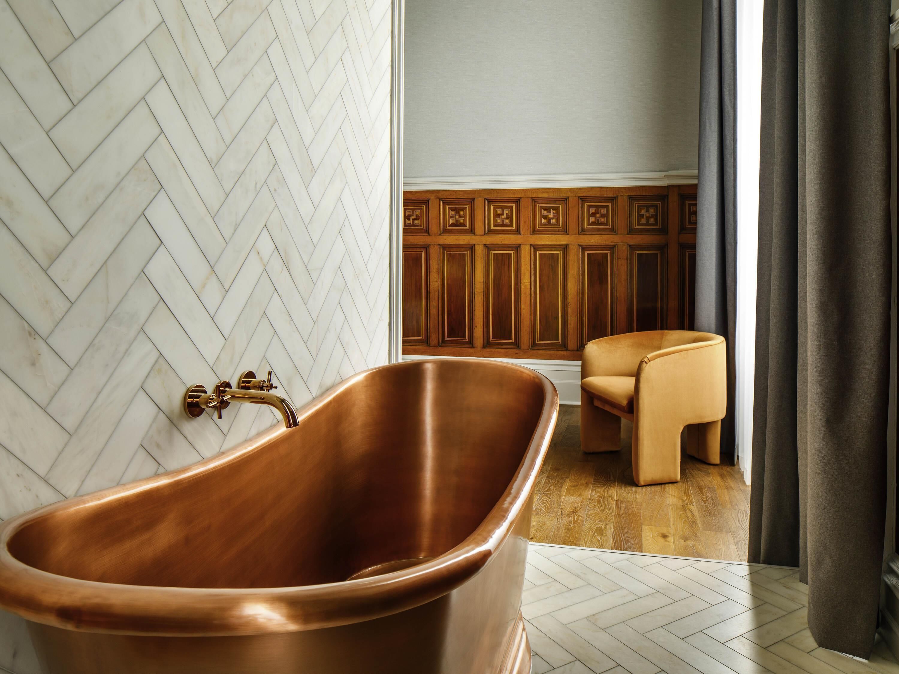 Ein Badezimmer mit einer Kupferbadewanne, Wänden mit Fischgrätmuster, Holzvertäfelung und einem Ledersessel.