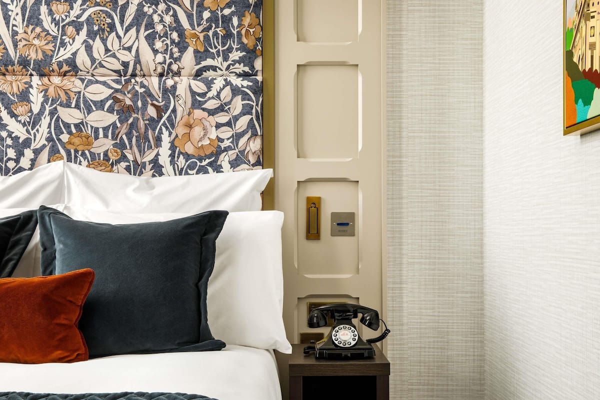 A hotel room with a bed, pillows, a nightstand with a telephone, and a door with a light switch.