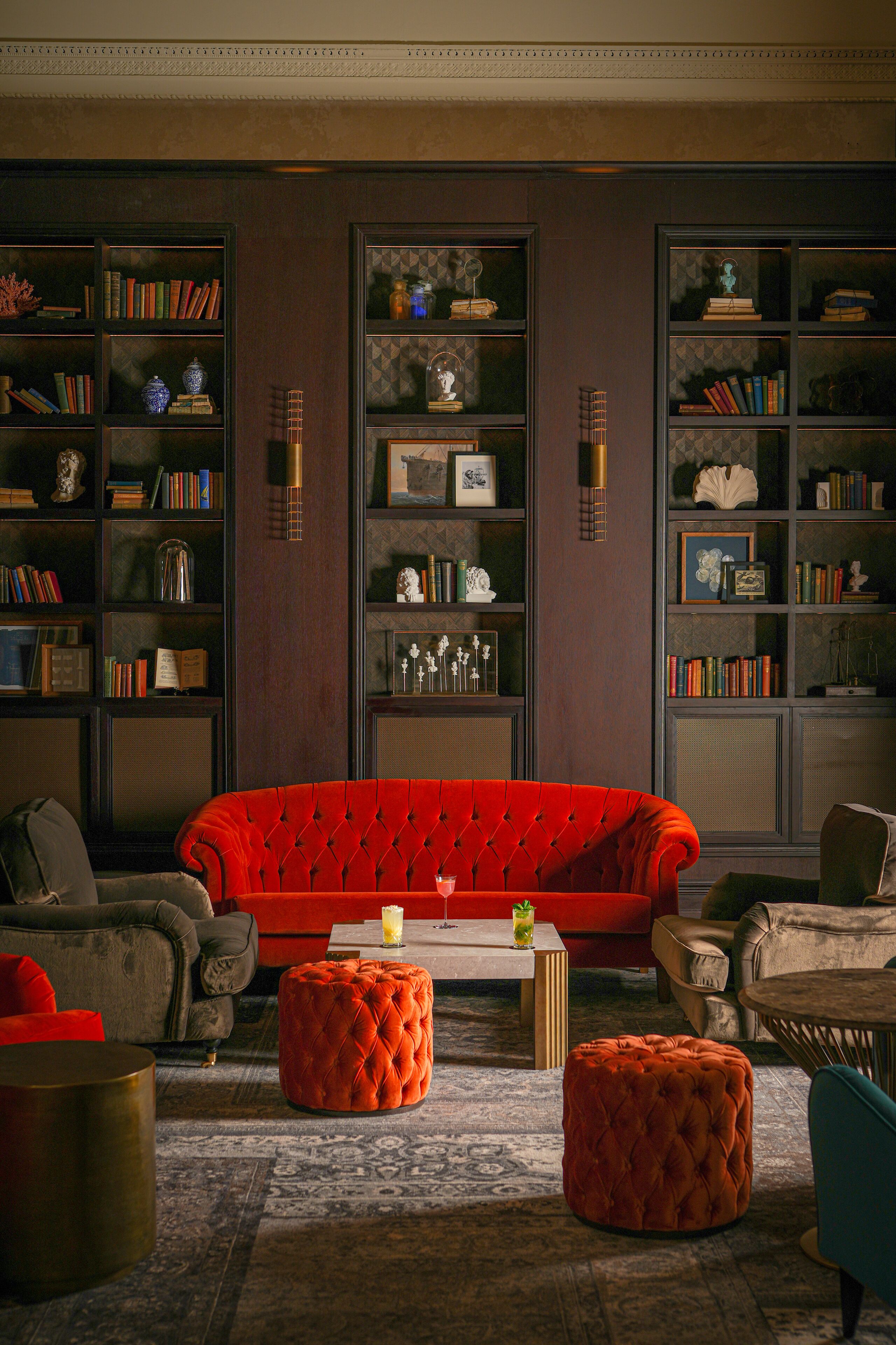 Una sala de estar con un sofá rojo, dos otomanas, una mesita de centro y una estantería llena de libros y objetos decorativos.