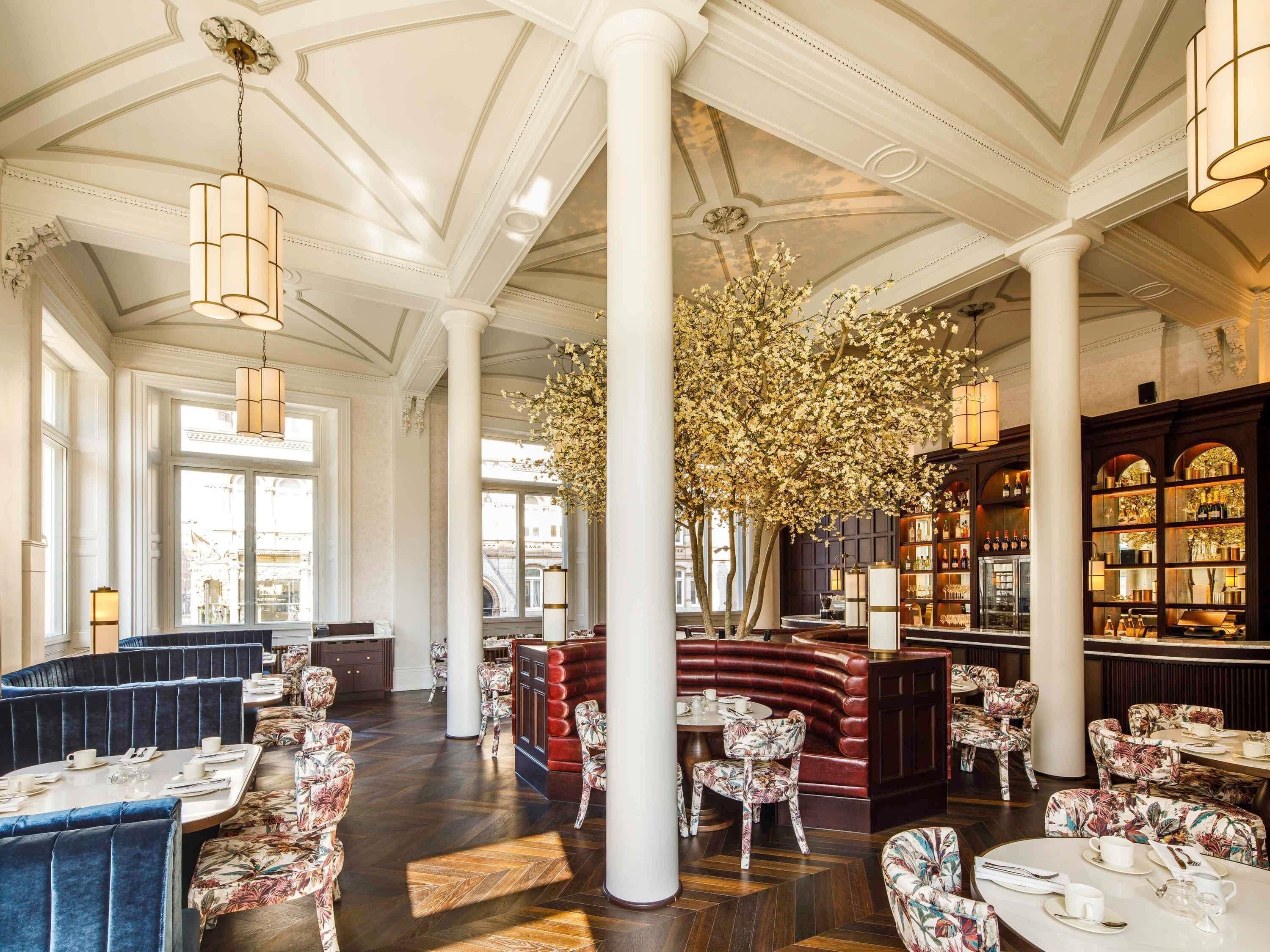 A spacious hotel lobby with high ceilings, large windows, and a central seating area with tables and chairs.