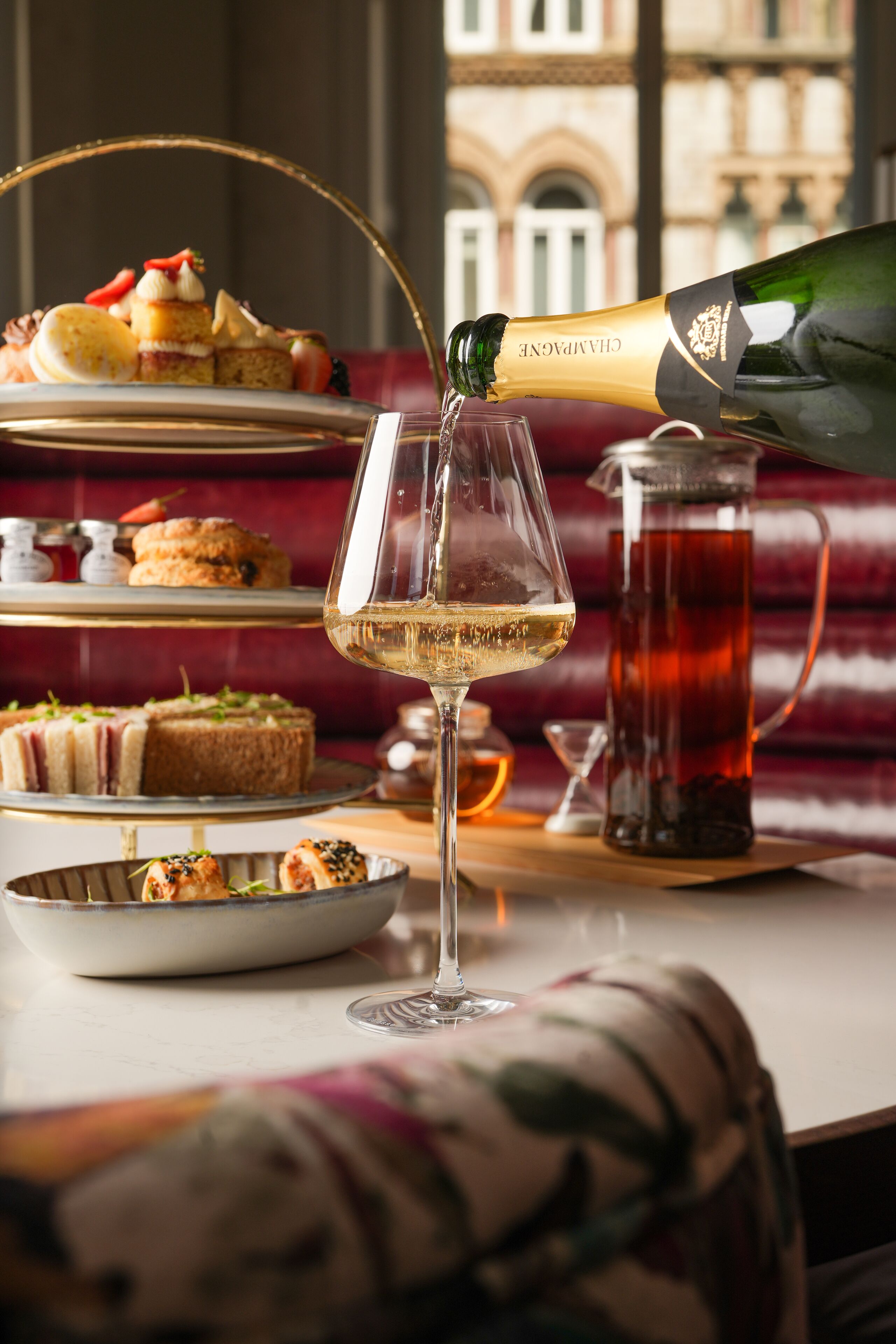 Une coupe de champagne est versée dans un verre à vin, avec une présentation de desserts en étages et une théière en arrière-plan.