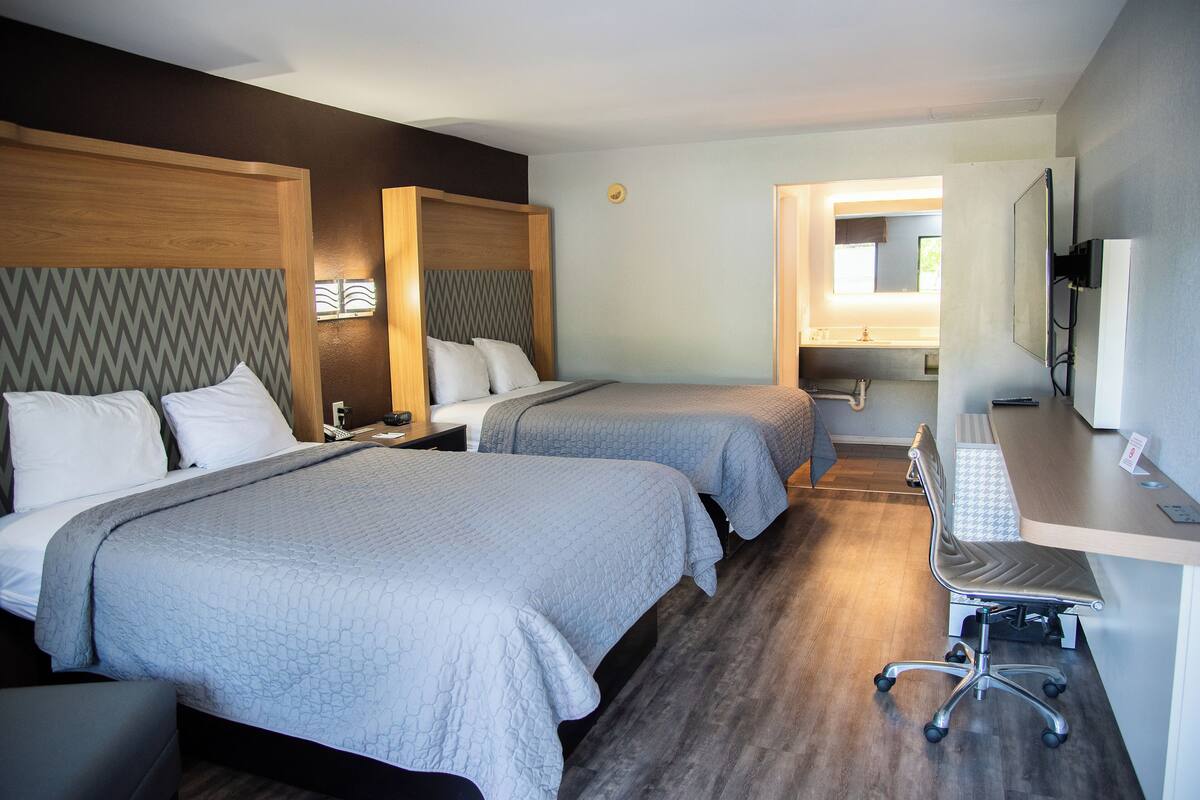 A hotel room with two beds, a desk, a chair, and a bathroom visible through an open door.