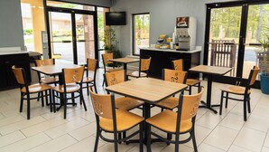 Un restaurante con varias mesas y sillas, un ventilador de techo, un televisor montado en la pared y vistas al exterior a través de amplias ventanas.
