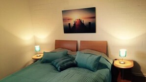 A neatly made bed with a green bedspread and pillows, two bedside lamps, and a sunset painting on the wall. - FeWo „Skipper“ 71m², 4 Pers. im Marinapark Scharmützelsee, Sauna, Whirlpool, Kamin (Wendisch Rietz Siedlung)